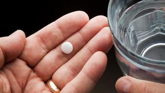 A hand holding an aspirin pill next to a glass of water