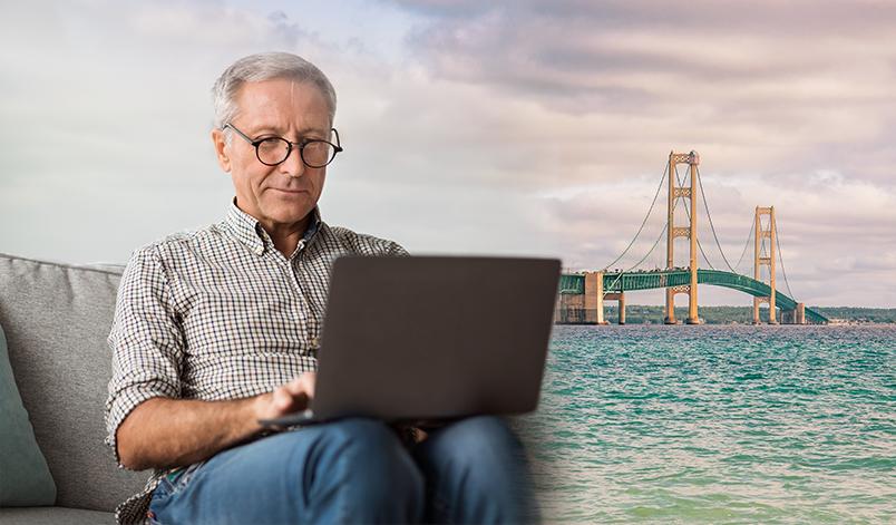 man sitting on the couch looking at laptop with background of Mackinaw Bridge